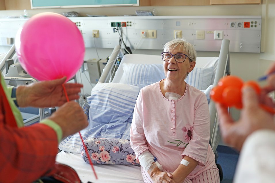 UKM | Eine Frau im rosafarbenen Schlafanzug sitzt auf einem Krankenbett und lächelt einen Clinik-Clown an. Im Vordergrund sieht man seine Hände, die einen pinkfarbenen Luftballon halten. Ein Moment der Freude im Klinikalltag.