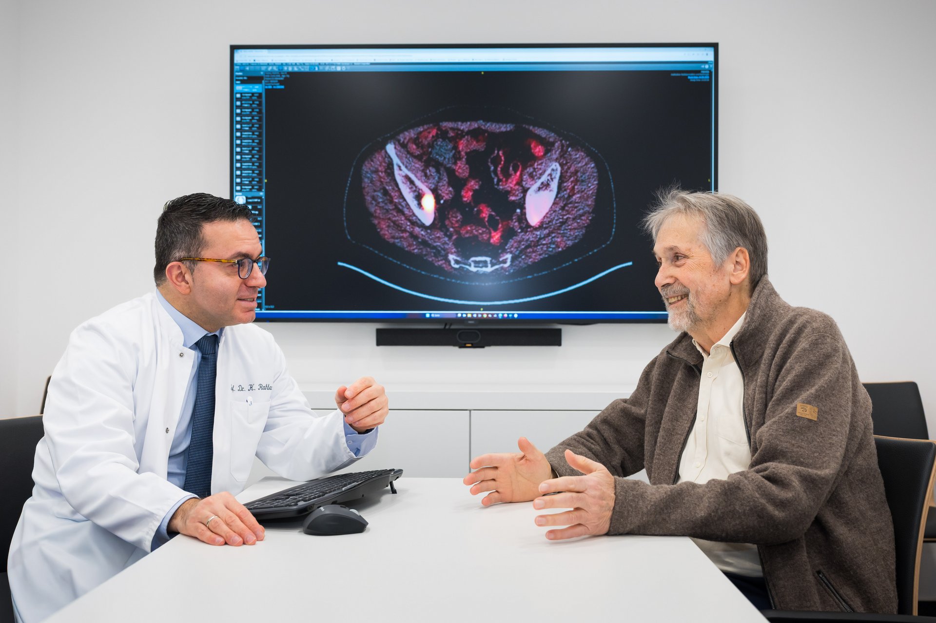 UKM | Arzt im weißen Kittel und Patient in Hemd und Jacke sitzen an einem weißen Tisch und sprechen. Im Hintergrund ein großer Monitor mit einer Bildgebung