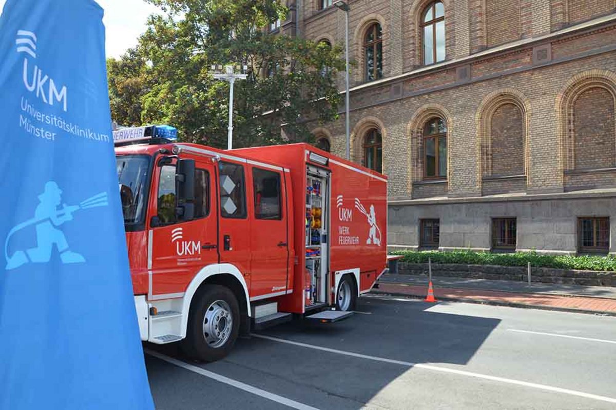 UKM Werkfeuerwehr | Ein Einsatzwagen ist von der Seite zu sehen. Vorne ist außerdem ein blauer UKM Werkfeuerwehr Banner. Im Hintergrund sind man ein Gebäude.