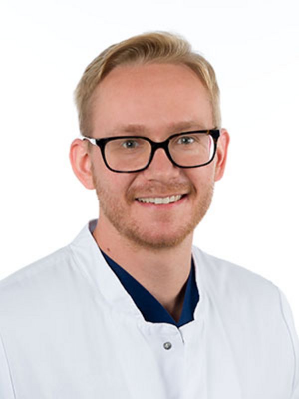 UKM Medizinische Klinik A | Lucas Brehm | Er trägt eine dunkle Brille, einen weißen Kittel und lächelt in die Kamera.