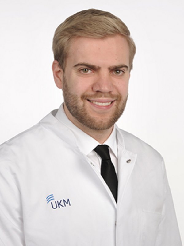 UKM Klinik für Strahlentherapie | Jan Siats | Er hat helle Haare, trägt eine Krawatte und einen Kasack und lächelt in die Kamera.