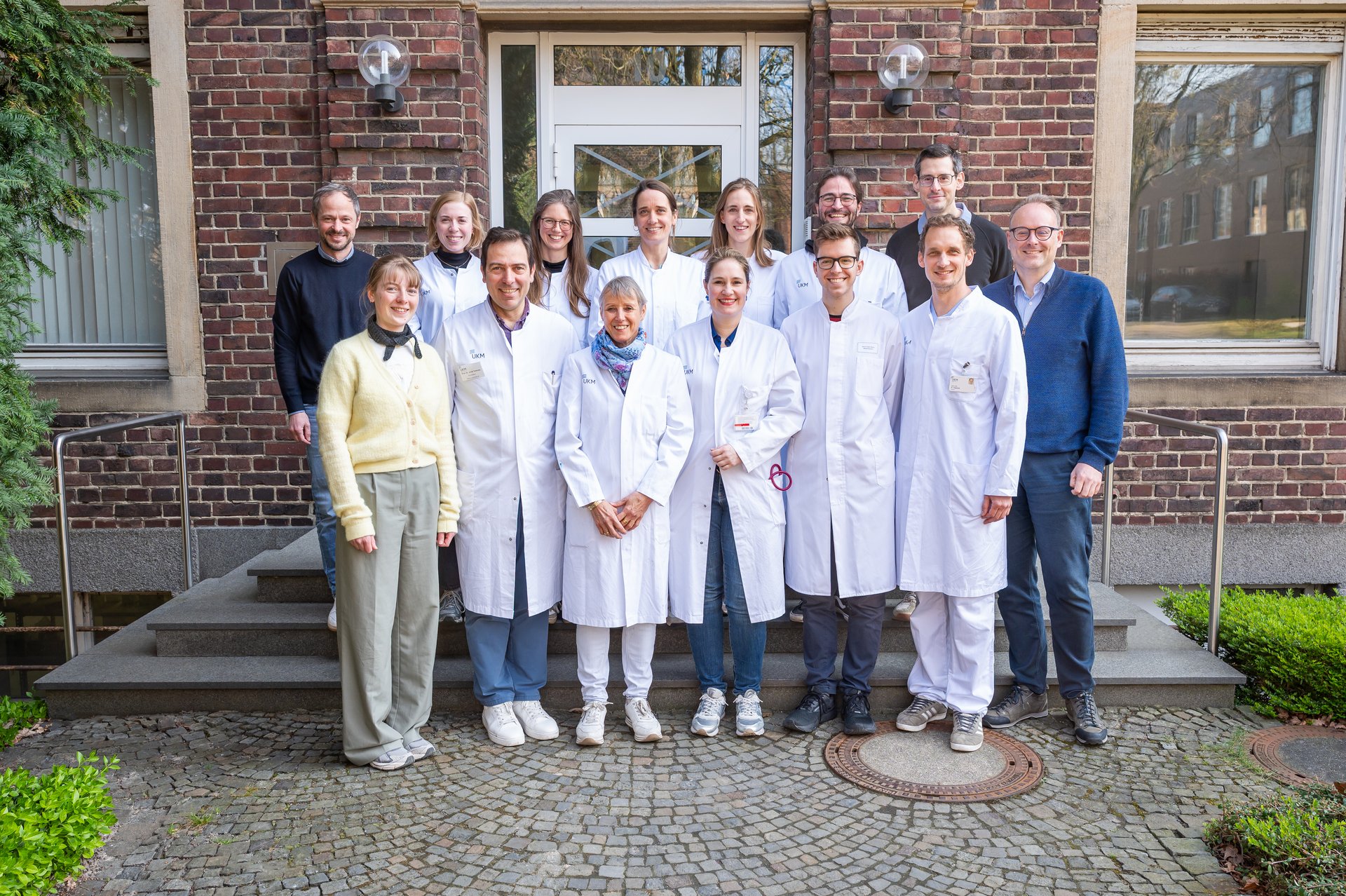 UKM | Interdisziplinäres Team mit 14 Personen. Sie stehen vor einem Klinkerbau in zwei Reihen und lachen; die meisten tragen einen weißen Kittel