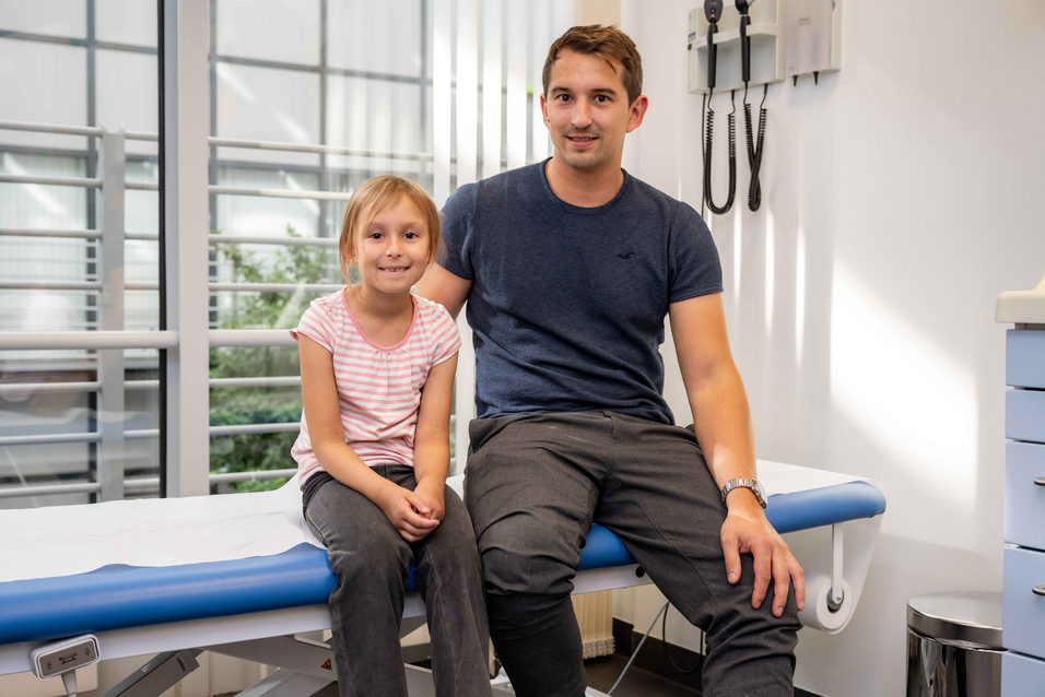 UKM | Die sechsjährige Rheuma-Patientin sitzt neben ihrem Vater auf einer Liege. Beide lächeln.