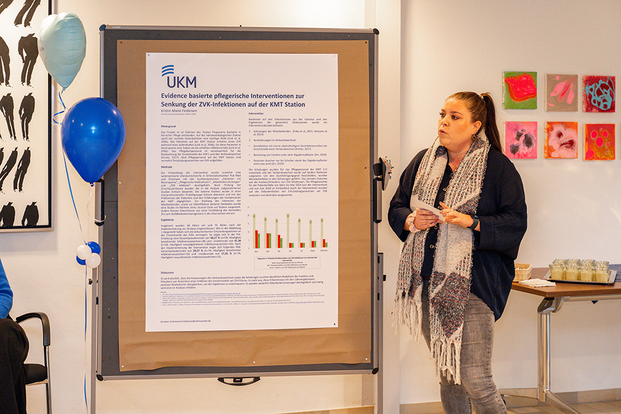 UKM |UKM-Veranstaltung | Eine Frau steht neben einem großen Poster zum Thema Evidenzbasierte pflegerische Interventionen zur Senkung der ZVK-Infektionen auf der KMT-Station