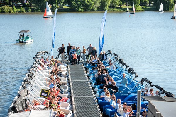 SchlauRaum | An einem Steg im Aasee liegen weiße Tretboote auf der linken Seite, blaue Tretboote auf der rechten Seite. In den Tretbooten sitzen Leute, sie winken fröhlich.
