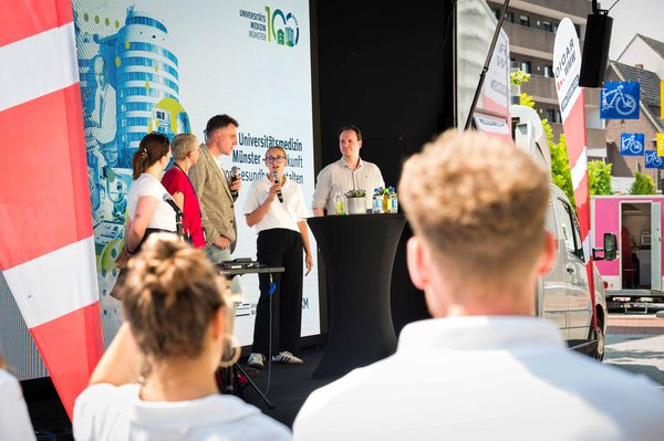 UKM | Vier Mitarbeitende des UKM stehen mit dem Moderator auf der Bühne. Im Vordergrund befinden sich Zuschauende.