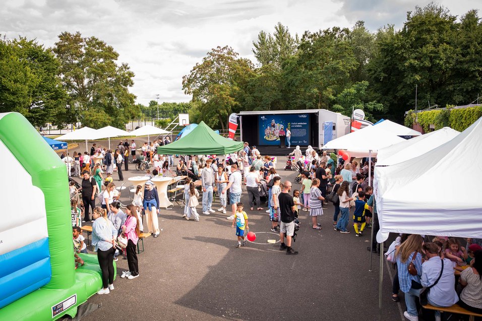 Asphaltierter Platz mit Stagetruck, weißen Pavillons und Sonnenschirmen, Hüpfburg und vielen Menschen. Im Hintergrund sind Bäume