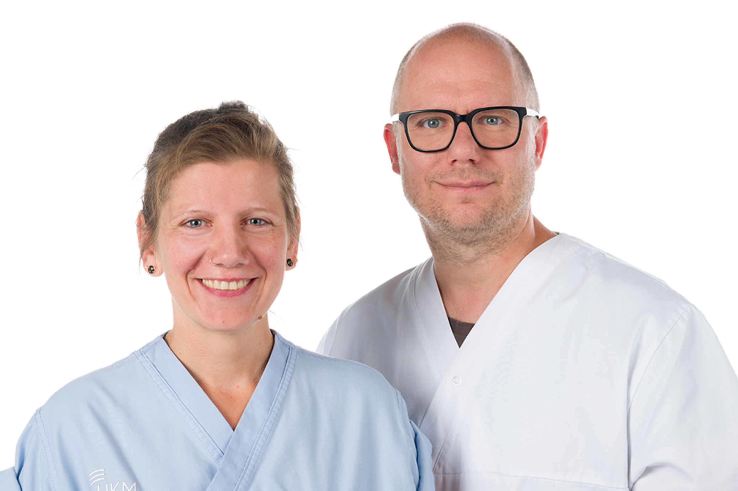 UKM | Portrait der Pflegekräfte Madeleine Barthmuß und Marc Bastin. Sie hat kurze blonde Haare und trägt einen hellblauen Kasack. Er trägt eine dunkle Brille und einen weißen Kasack. Beide lachen.