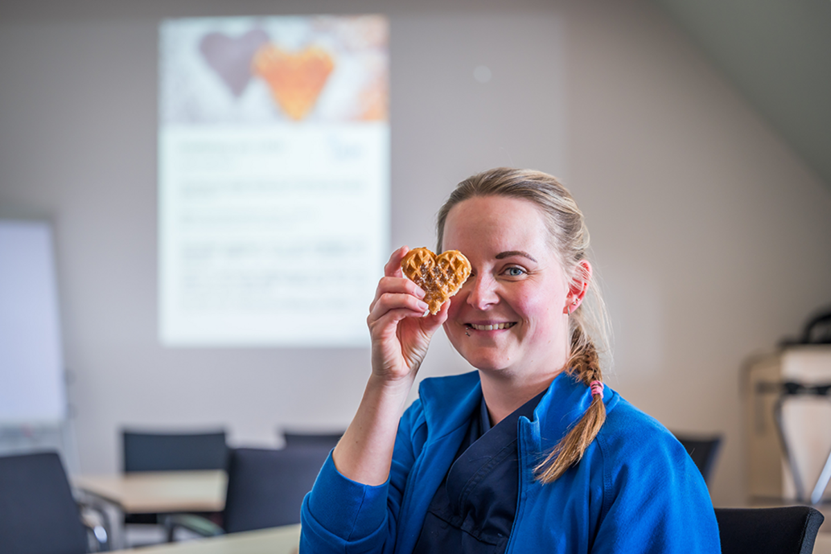 UKM | Mitarbeiterin aus der Allgemeinchiurgie hält sich eine herzförmige Waffel vor ein Auge und lacht. Im Hintergrund ein Poster mit zwei Herzen