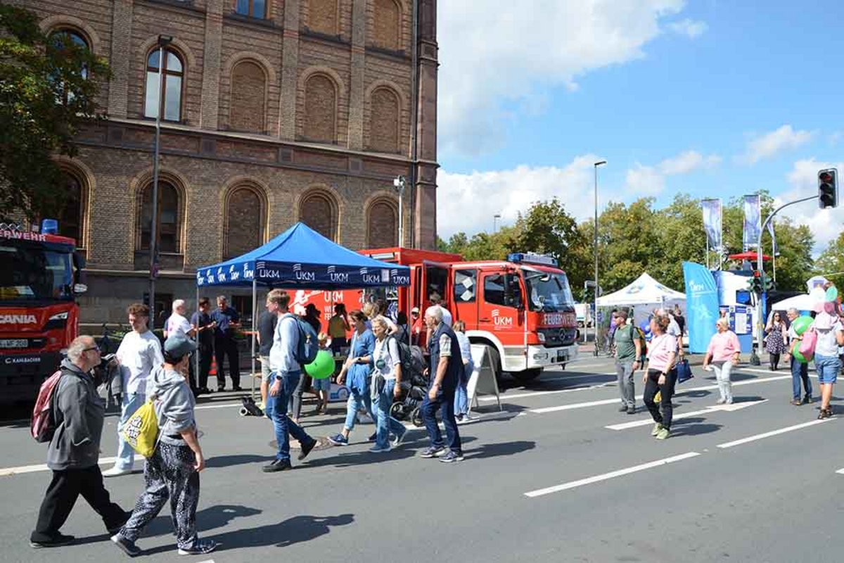 UKM Werkfeuerwehr | Man sieht ein Feuerwehrwagen von weiter weg. Davor sind viele Menschen auf der Straße unterwegs.