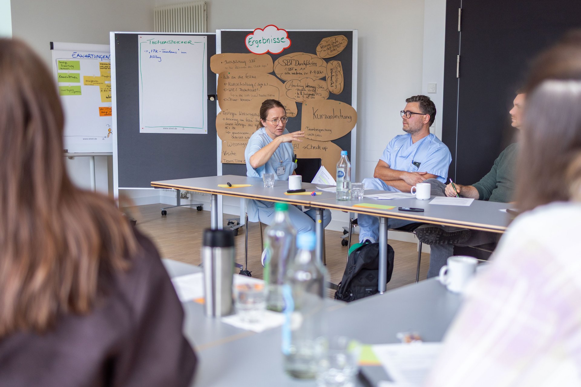UKM Magnet | Eine Frau im Kasack im Fokus, sie gestikuliert. Um sie herum sitzen weitere Menschen und hören ihr zu. 