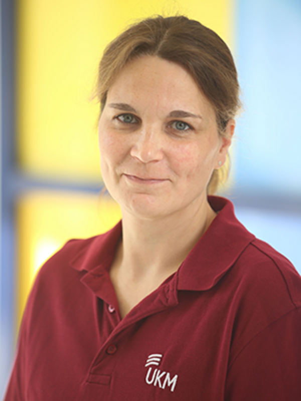 UKM | Portrait einer Mitarbeiterin aus der Palliativmedizin. Sie trägt einen dunkelrotes Poloshirt und lächelt. Blau-gelber Hintergrund
