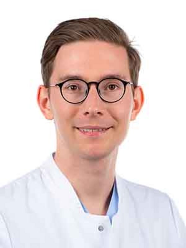 UKM Medizinische Klinik B | Fabian Schachteli | Er trägt eine Brille und einen weißen Kittel und lächelt in die Kamera.