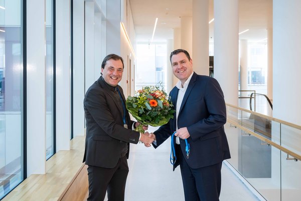 UKM | Zwei Männer in dunklen Anzügen geben sich die Hand und lachen. Der eine überreicht dem anderen einen Blumenstrauß. 
