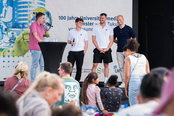 Drei UKM-Mitarbeiter stehenbei der UKM-Roadshow auf der Bühne und lachen. Links steht der Moderator und spricht ins Mikrofon. Im Vordergrund ist Publikum zu sehen. Im Hintergrund ein großes Banner zum Jubiläum 100 Jahre Universitätsmedizin Münster