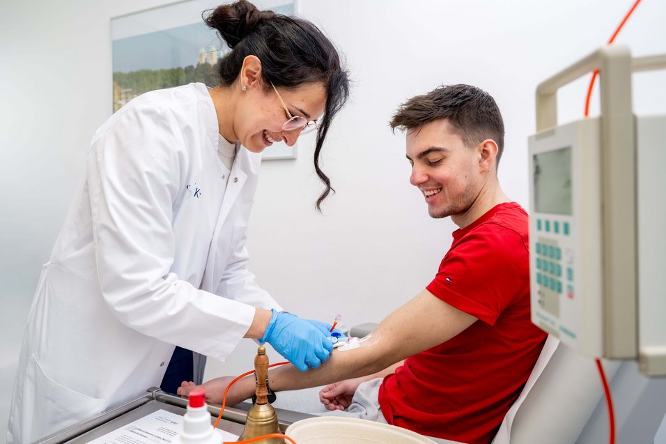 UKM | Ärztin im weißen Kittel und mit blauen Handschuhen schließt den Lupus-Patienten über einen Portkatheter am Arm an die Infusion an. 