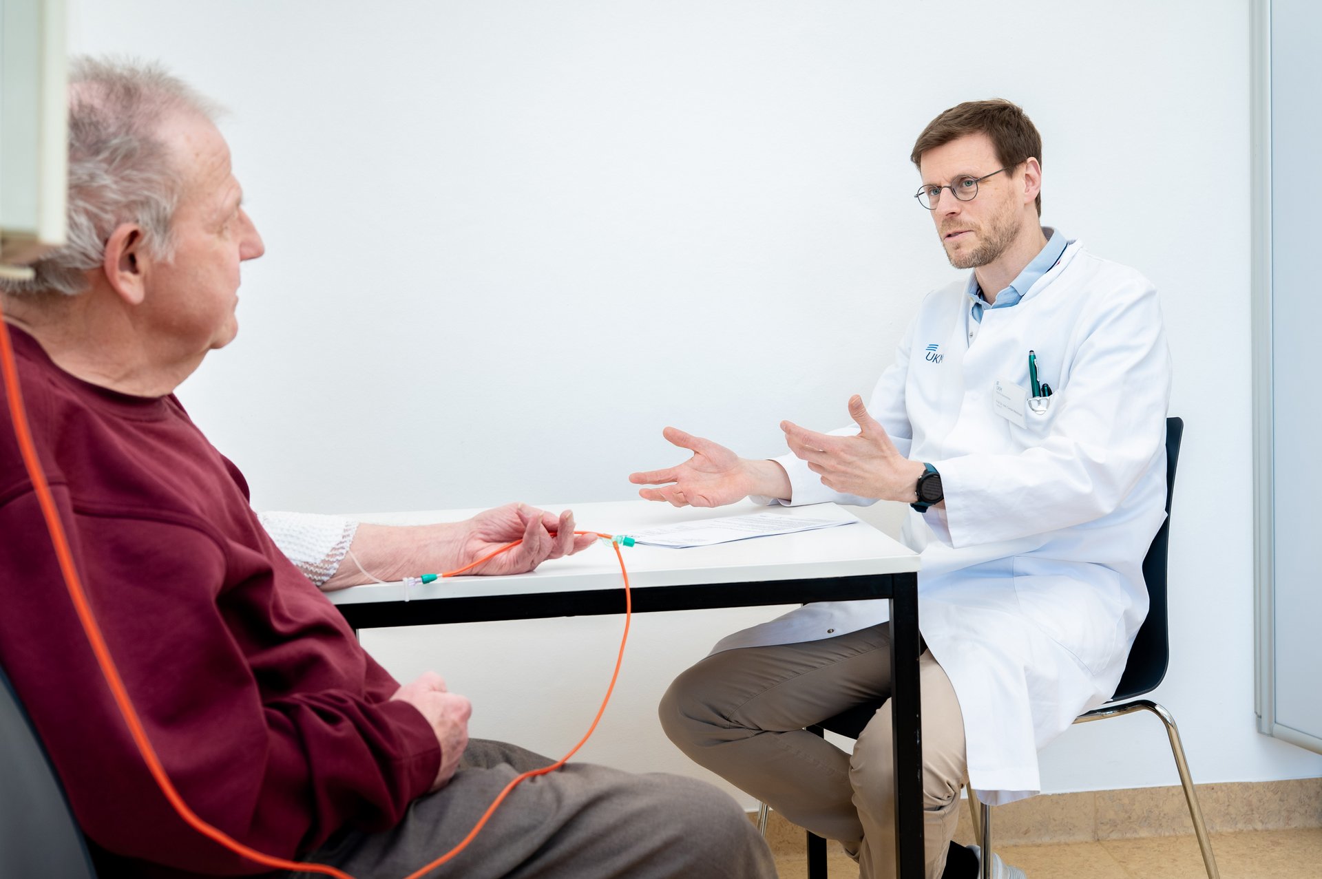 UKM | Arzt im weißen Kittel und Patient sitzen an einem Tisch.