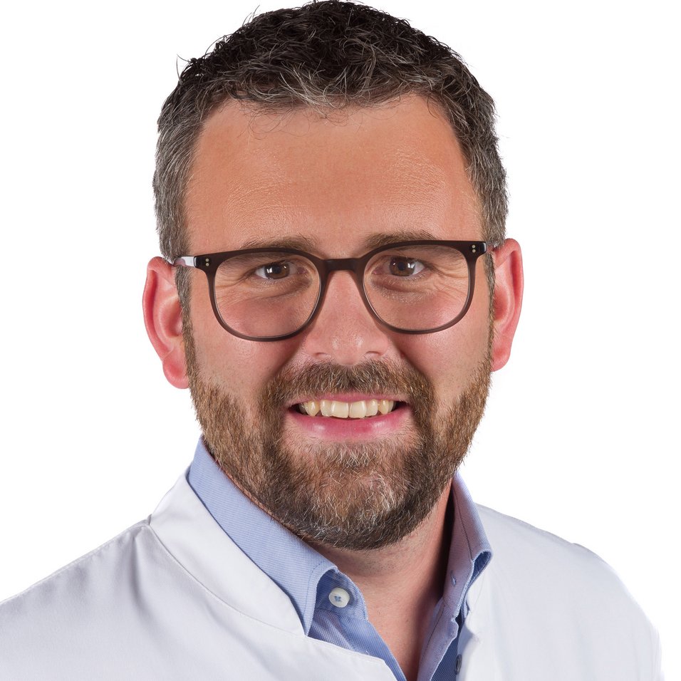 UKM | Portraitaufnahme von Unfallchirurg Simon Öckenpöhler. Er trägt einen weißen Kittel, eine Brille und lächelt.