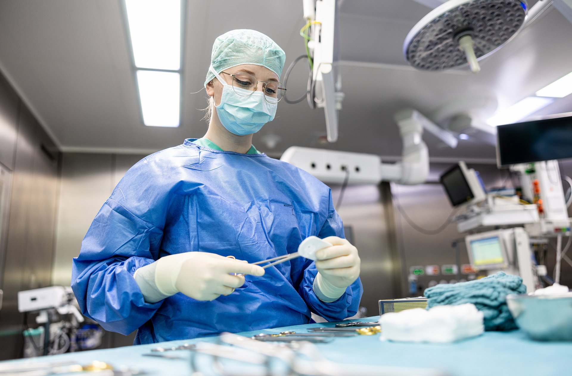 OP-Mitarbeiterin in Schutzkleidung im Operationssaal; sie hält eine Zange in der Hand