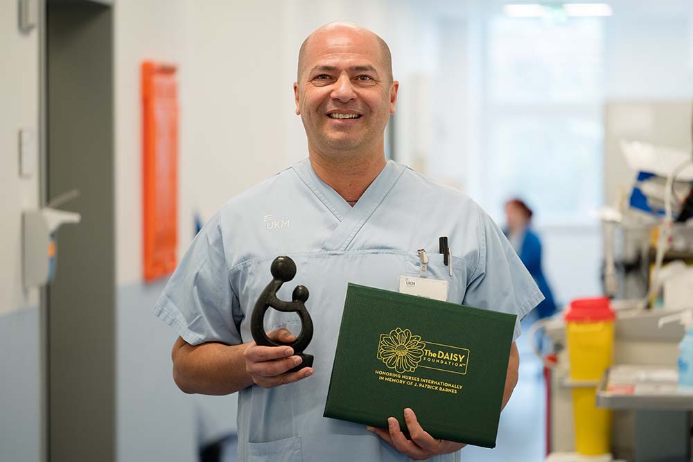 UKM Daisy Award | Portraitbild von Flavcio de Oliveira. Er lächelt in die Kamera und hält den Preis und die Urkunde in den Händen.