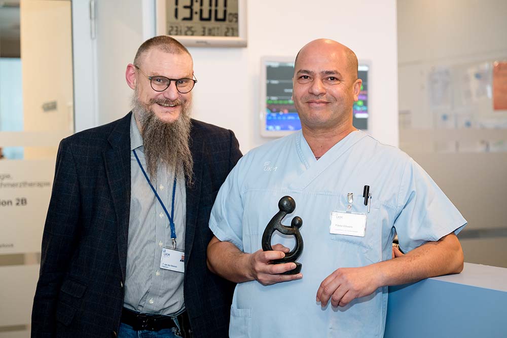 UKM Daisy Award | Zwei Männer stehen nebeneinander und lächeln in die Kamera. Links steht Thomas van den Hooven. Rechts neben ihm steht Flavio de Oliveira und hält den Daisy Award in der Hand.