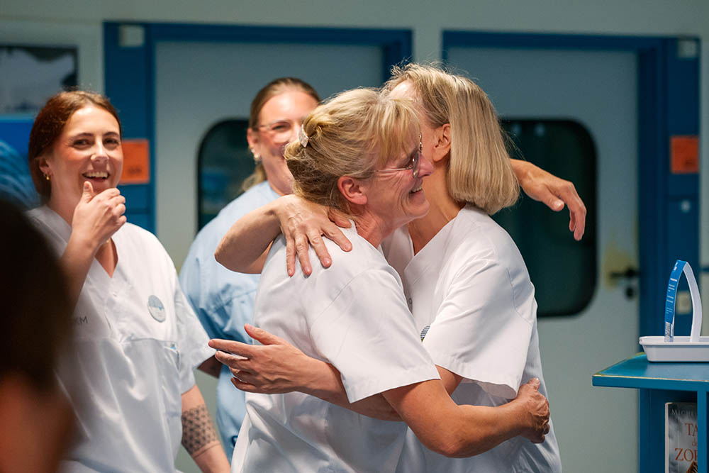 UKM Daisy Award | Andrea Kümper umarmt eine Kollegin auf der Station. Um sie herum stehen weitere Mitarbeitende und freuen sich. 