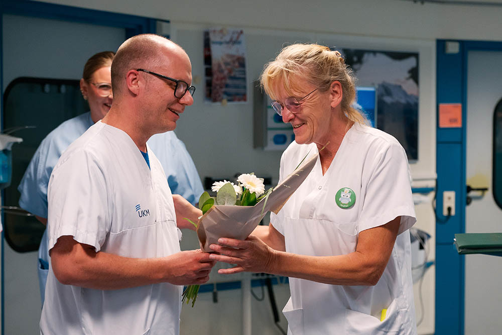 UKM Daisy Award | Ein männlicher Kollege im weißen Kasack überreicht Andrea Krümpel Blumen und einen Umschlag.
