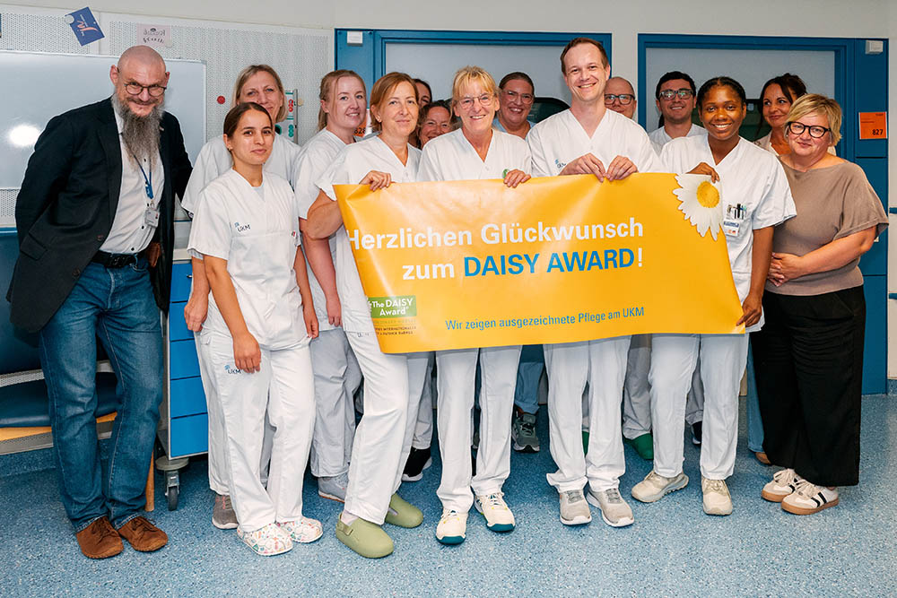 UKM Daisy Award | Ein Gruppenbild mit Mitarbeitenden der Station. Sie halten einen gelben Daisy Award Banner in den Händen und lächeln alle in die Kamera.