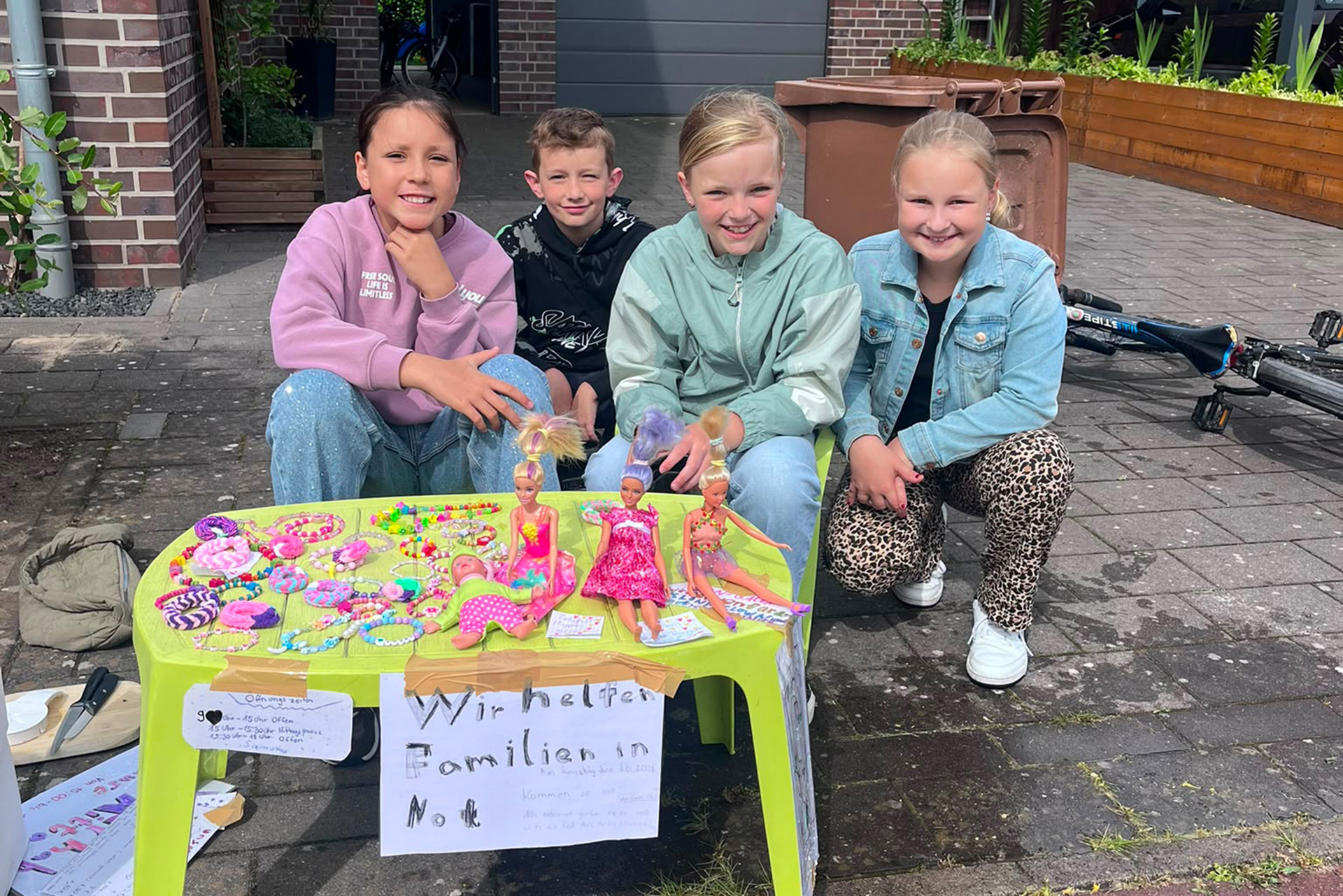 UKM Spendenmitteilung | Vier Kinder hocken vor einem kleinen Tisch. Auf diesem liegen die selbstgeknüpften Armbänder. Davor hängt ein Schild mit der Aufschrift "Wir helfen Familien in Not"